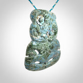 This photo shows a hand carved Aotea stone Tiki pendant. It is a very light blue colour with pāua shell insert eyes. The cord is blue and silver grey and is length adjustable